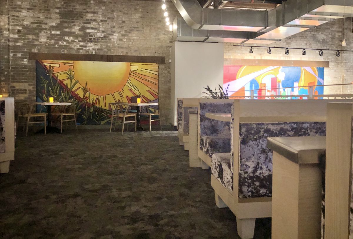 Banquettes upholstered with lavender velvet line the right side of the lofted space. A red, yellow, and orange sun mural dominates the brick wall at the center, back of the room. Two two top tables are placed in front of it.