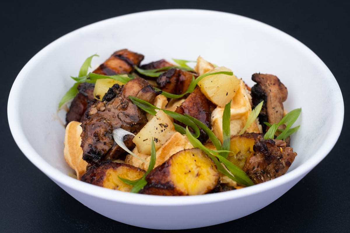 A white bowl filled with charred jerk chicken pieces, slices of golden plantains, and garnished with slices of green onions