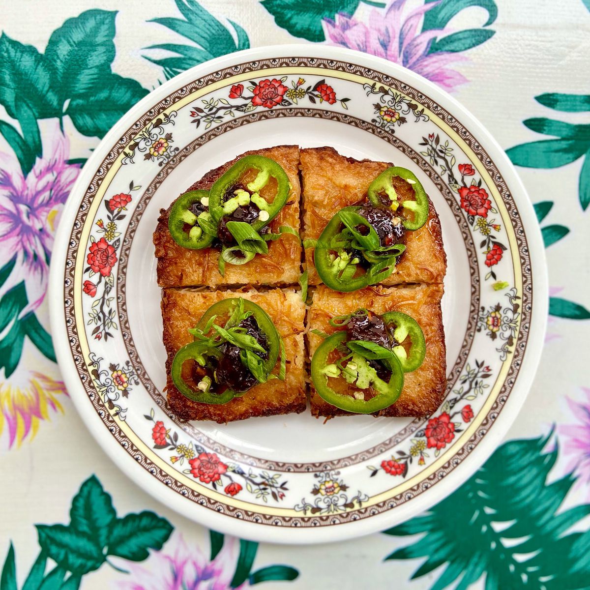 A large square of shrimp toast topped with large rings of jalapeño peppers on a China dish with a brightly colored tablecloth background