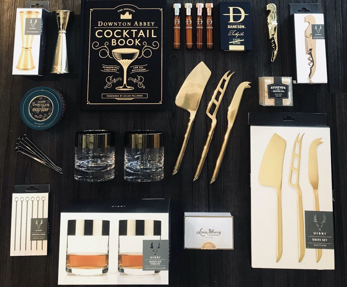 On a dark background are all the things that come in the kit, including gold cheese knives, lowball glasses with a silver rim, cocktail garnish skewers, gold corkscrew, gold shot glass, and more