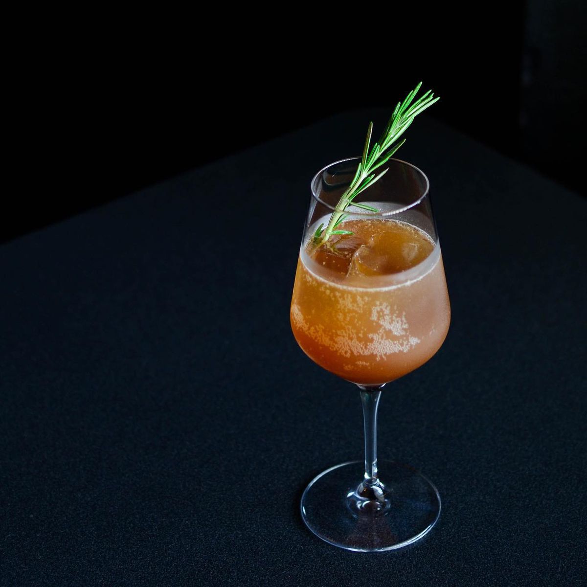A chilled orange beverage over a big ice cub with a rosemary garnish in a wine glass on a black topped bar