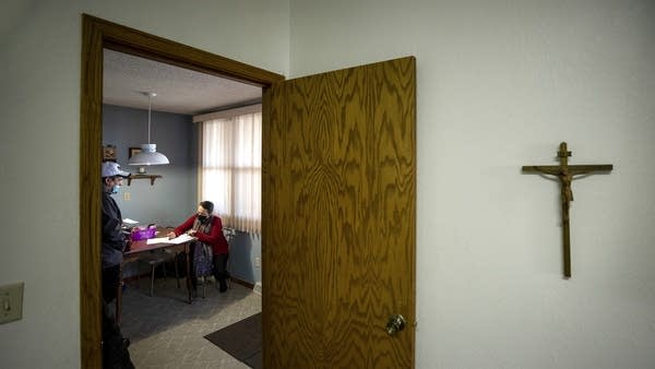 A cross hangs on the wall outside a room where a person sits at a table. 