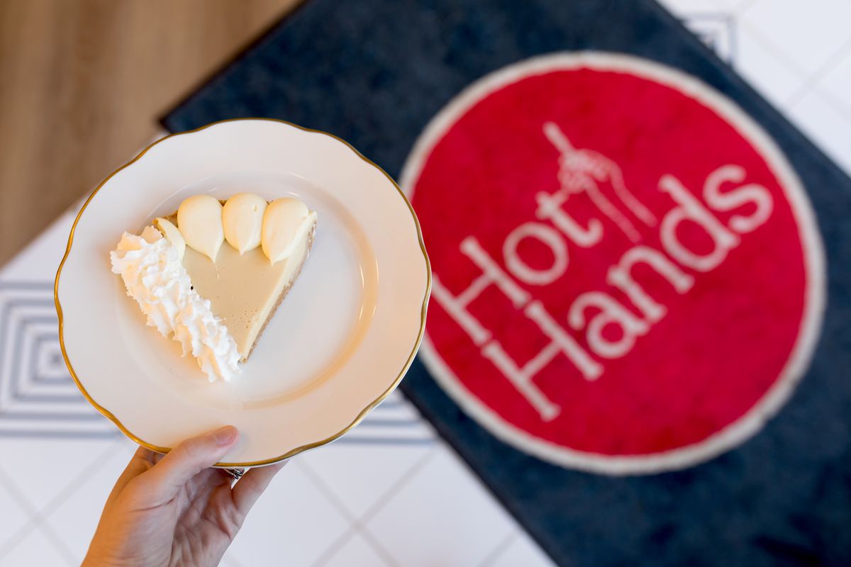 A hand holds a slice of vanilla creme pie over the tiled floor and Hot Hands logo on a rug below