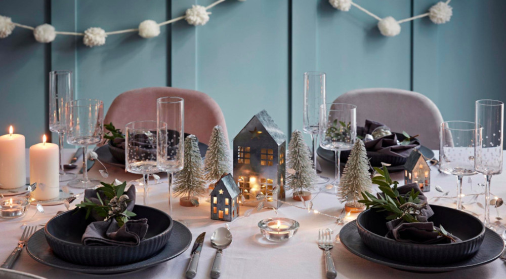 A table inside Stewart’s set with black plates on a white tablecloth. There are candles and small houses next to bottle brush green trees dusted with fake snow