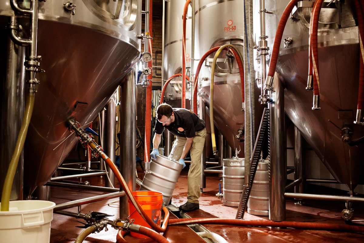 A brewer surrounded by tanks lifts a filled keg