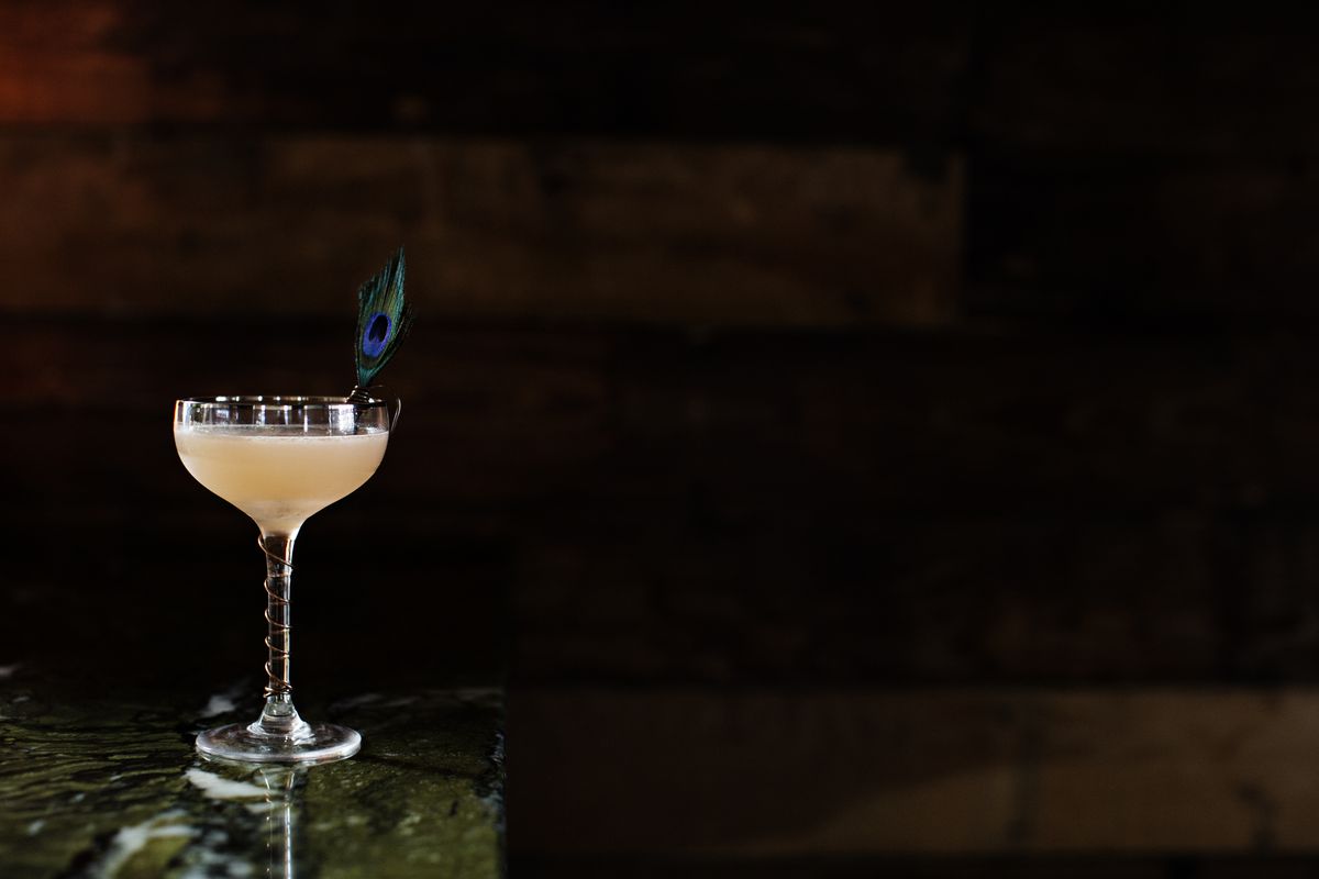 A striking image of the Naked Ballerina cocktail at Martina. A pink beverage in a coup glass is garnished with a peacock feather in front of a darkened wood wall on a green stone bar top