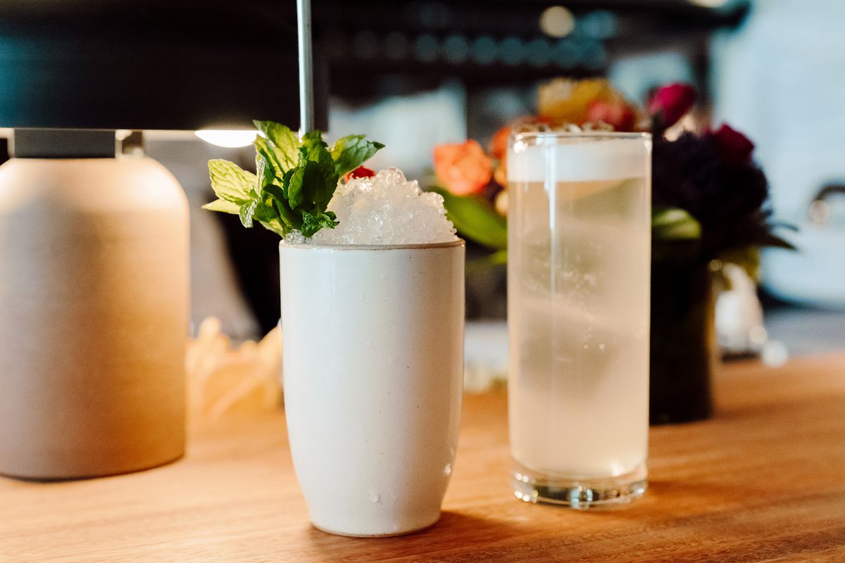 Two cocktails are pictured. One in an opaque eartenware tall glass, topped with a mound of pebble ice, a metal straw, and a mint bunch garnish. The other in a typical collins glass has an ever-so-slightly cloudy appearance with a thin layer of egg white froth on the top.
