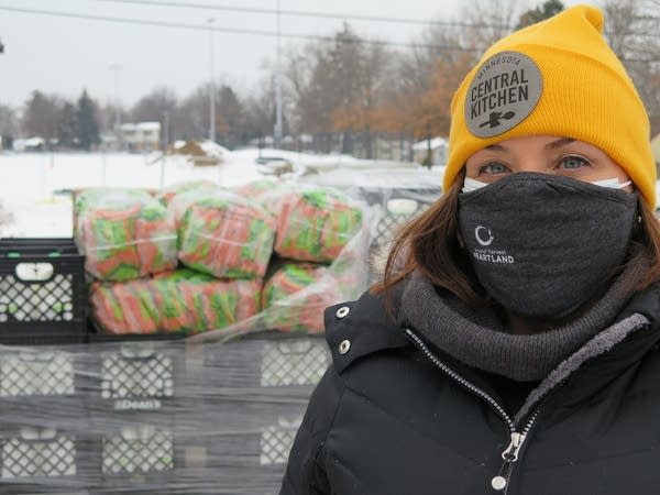 A woman stands outside with a face covering and a yellow winter hat.