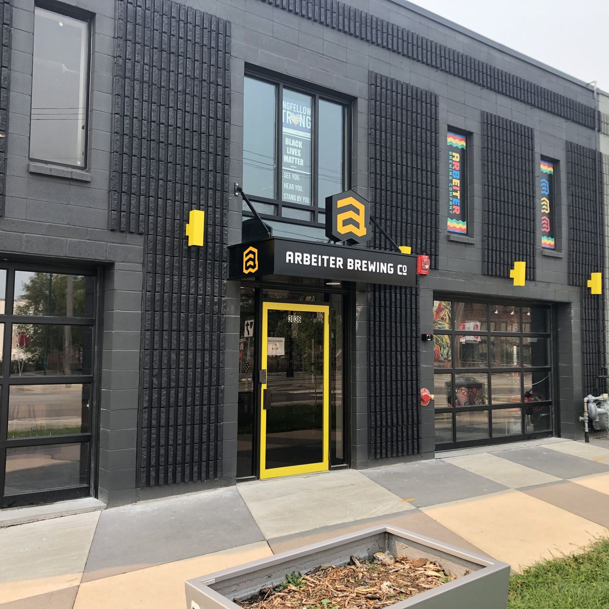 The black painted concrete walls are offset by bright yellow signage for the brewery. There are large windows facing the street.