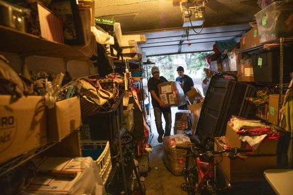 Two men carry boxes into a packed garage.