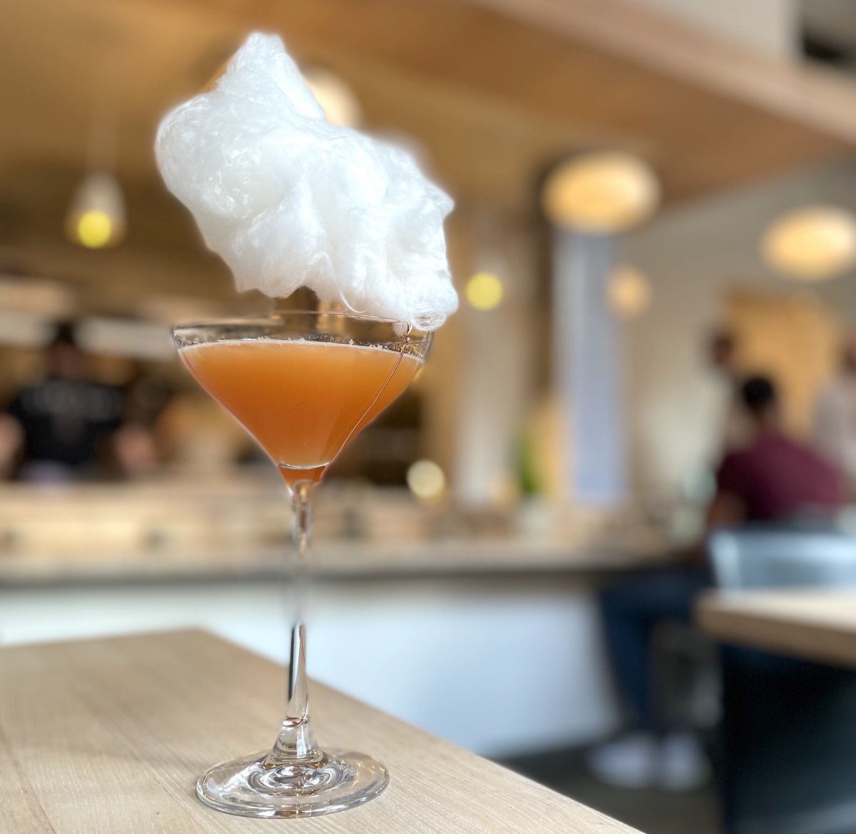 A martini glass is filled with orange liquid on a blonde wood table. It appears that there is a floating cloud above the glass