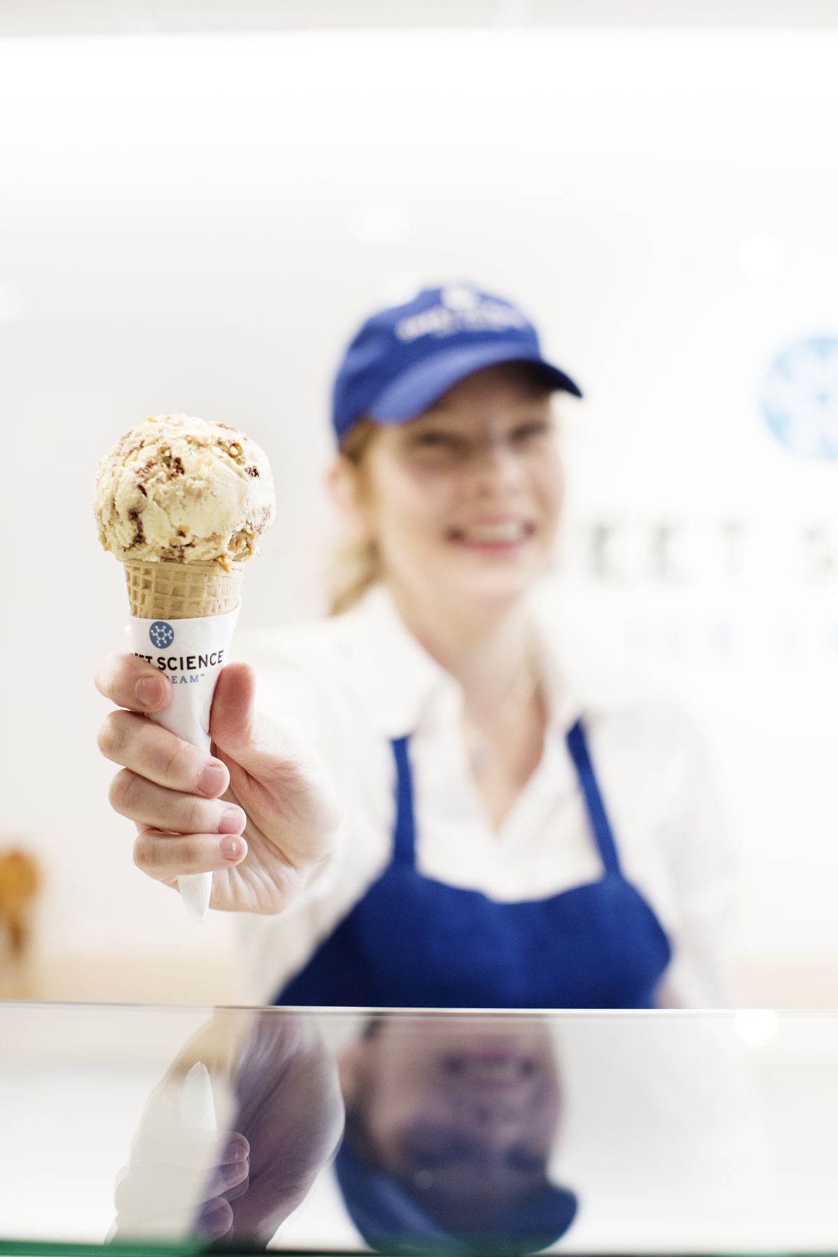 A smiling redhead in a blue Sweet Science shirt and apron, wearing a white shirt, is blurred behind the in-focus ice cream cone she’s holding. It’s a sugar cone with a giant vanilla-colored scoop studded with what looks like caramel pieces
