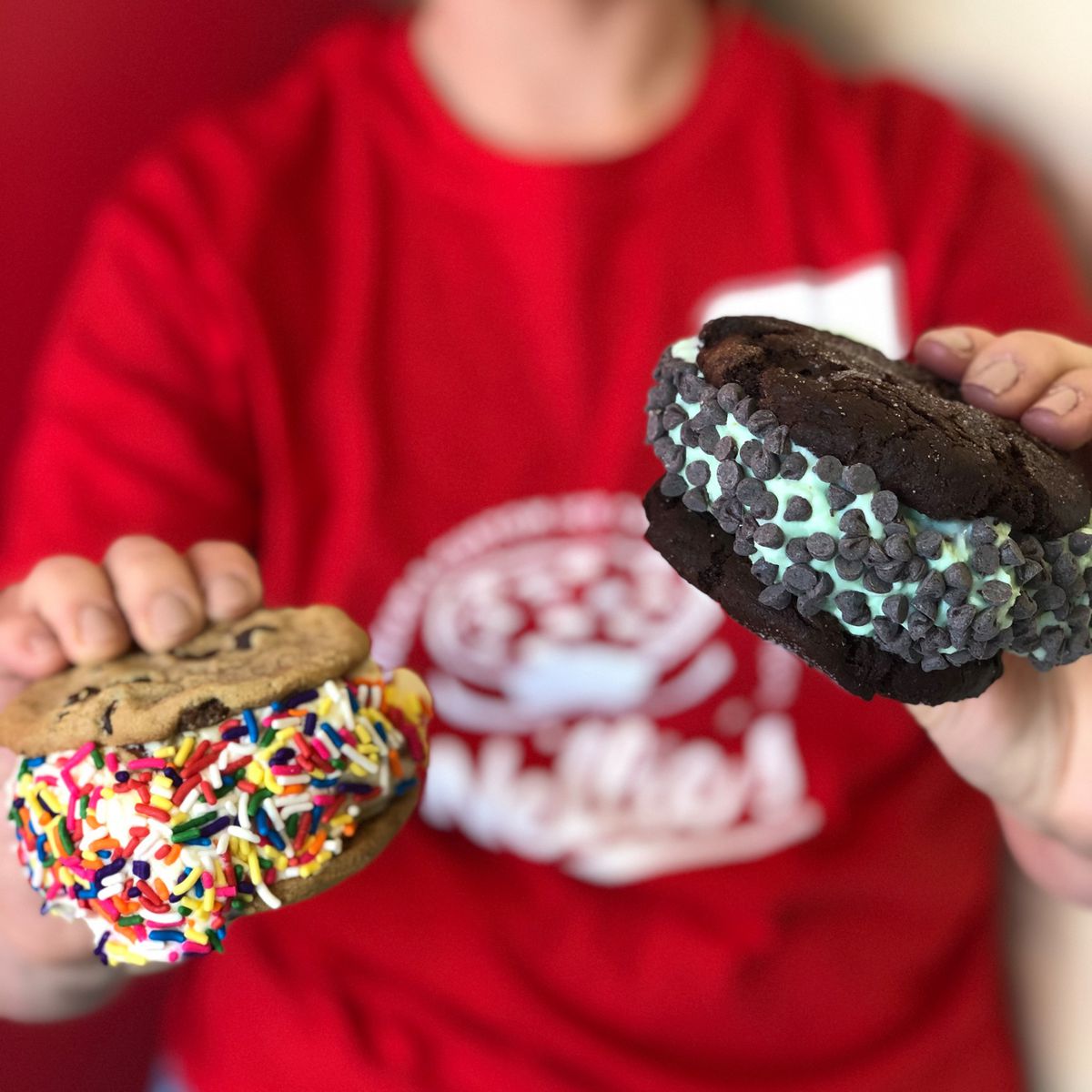 A worker in a red shirt holds two ice cream sandwiches, one with chocolate chips and the other with rainbow sprinkles