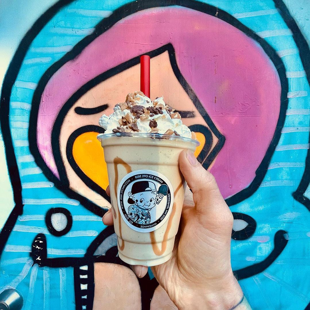 A hand holds a shake in a clear container with a proudly tattooed naked baby cartoon sticker on it in front of a brightly colored mural. Inside the shake are visible caramel drizzles and on top is a swirl of whipped cream and a bunch of candy chunks.