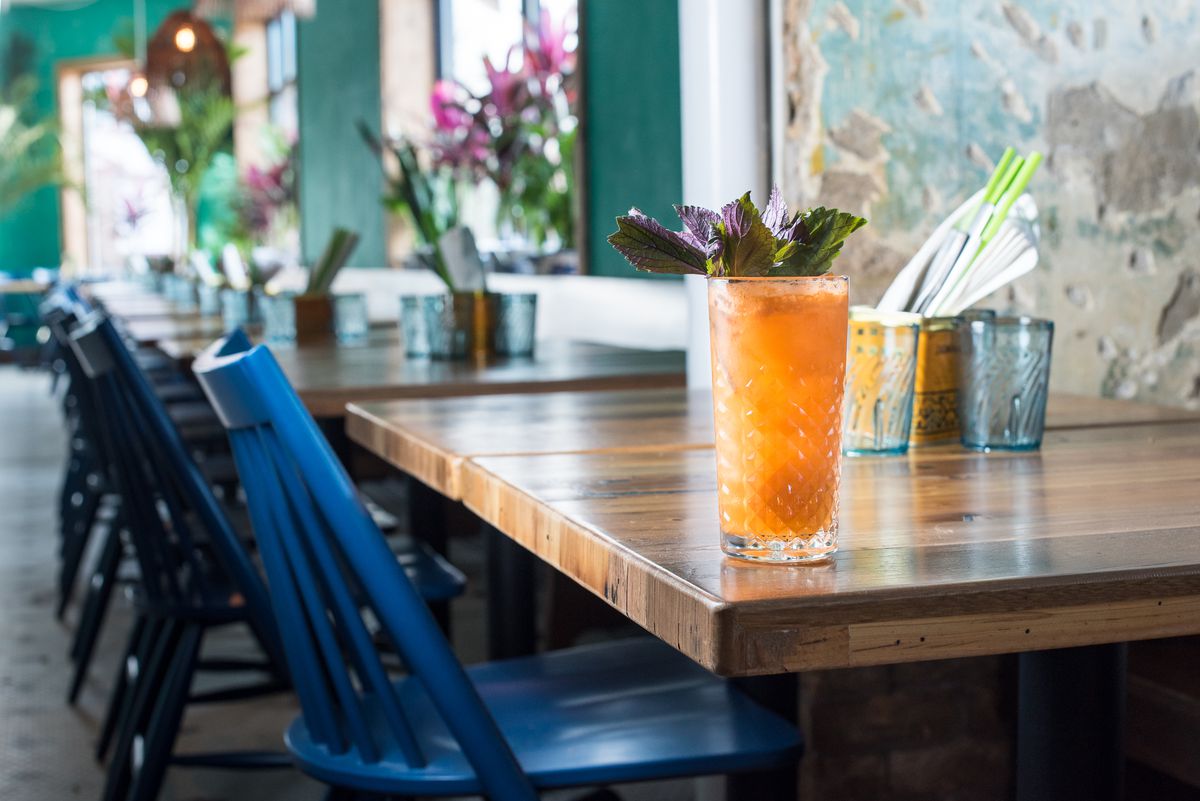 A cocktail in a tall glass with orange Thai tea is garnished with a bouquet of purple basil. It sits on a wooden table top. The vibrant turquoise and green tropical tones of the restaurant are visible in the background