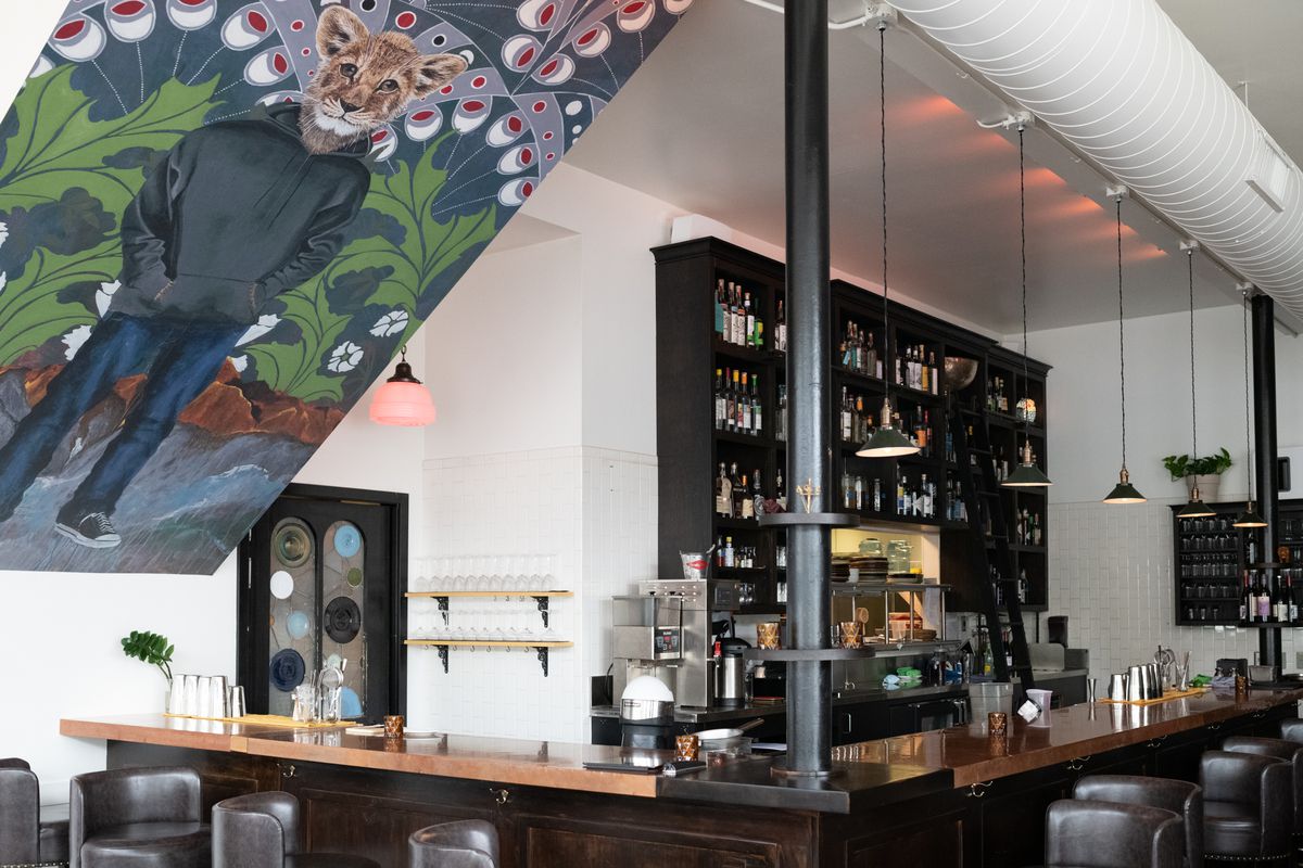 The interior of the restaurant with a side view of the bar. At the front, on the ceiling is a mural of an adolescent with a lion head and human body, in a zip up sweatshirt, jeans, and sneaks. The background is an abstract flower pattern in blue, white, and red.
