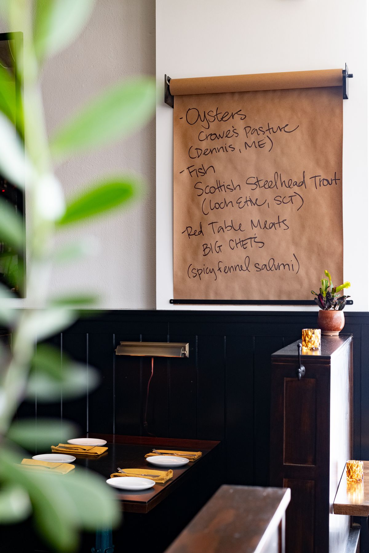 A dark brown wood booth set for four with bright, mustard-colored cloth napkins. Above it is a butcher paper specials listing the variety of mussels, fish, and salumi offered that day.