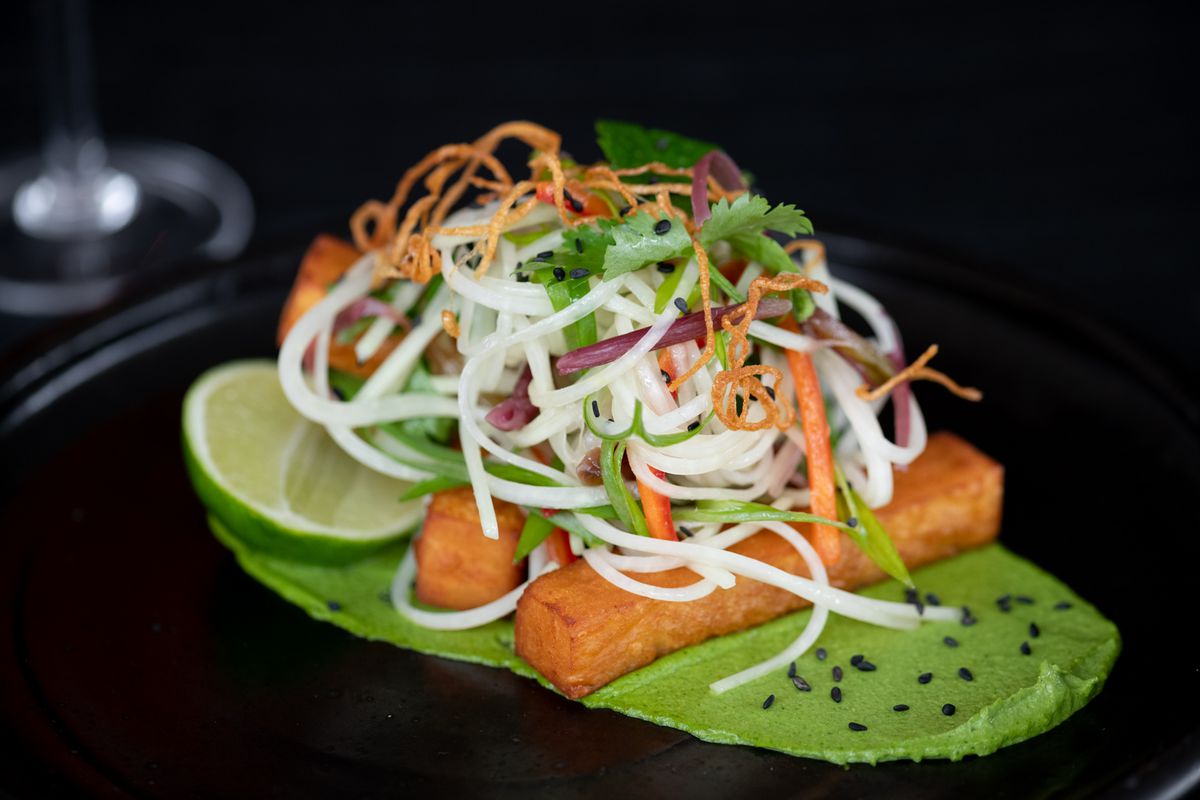 Two perfectly rectangular wedges of fried masa dough are stacked on top of a thick green sauce swiped on the black plate. On top are tenderils of rice noodles, micro cilantro, shaved carrot whisps and a smattering of black sesame seeds.