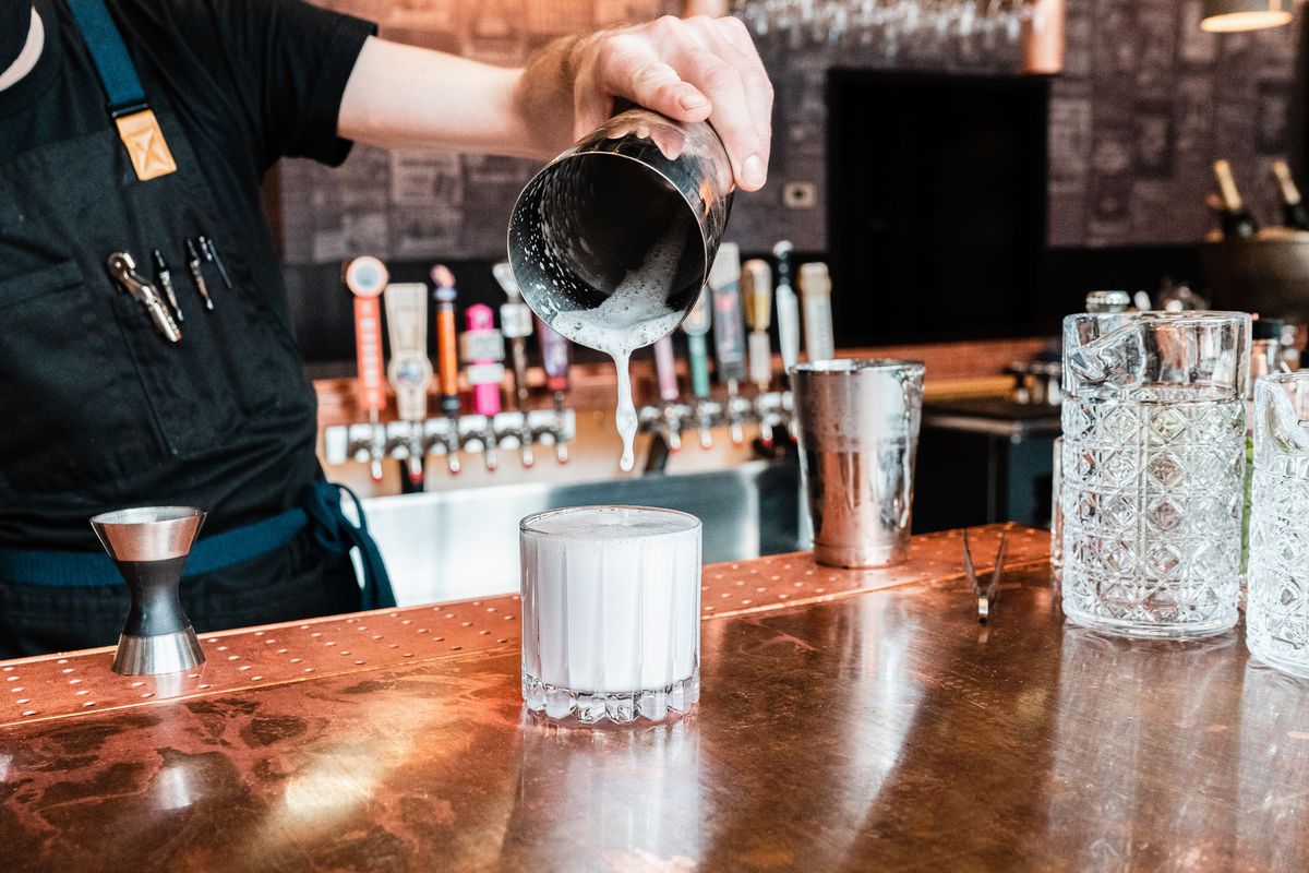 A bartender pours a frothy drink into a lowball glass from a silver tin shaker. 