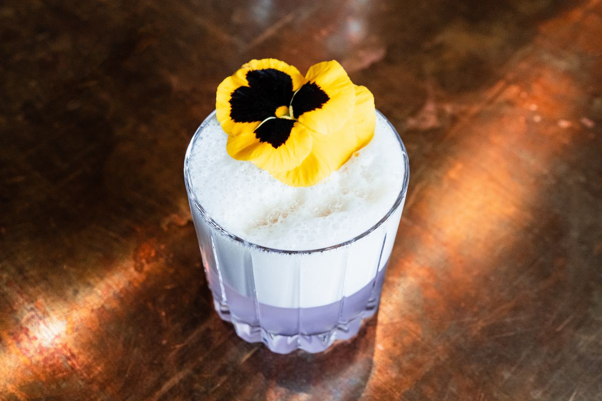 A drink with a frothy top is garnished with a yellow pansy. The liquid inside is a pale violet color.