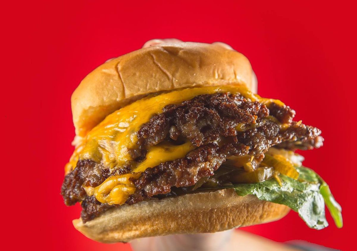A hand holds a double stacked, crusty burger with melted orange cheese on two buns with a bit of lettuce sticking out of the bottom on a red background.