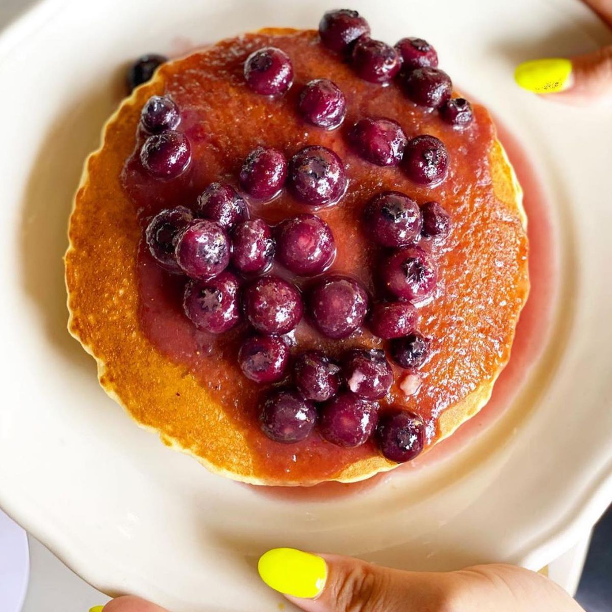 Two hands with a bright yellow manicure holds a white plate with a toasty colored pancake drenched in macerated cherries and a dark, sticky syrup