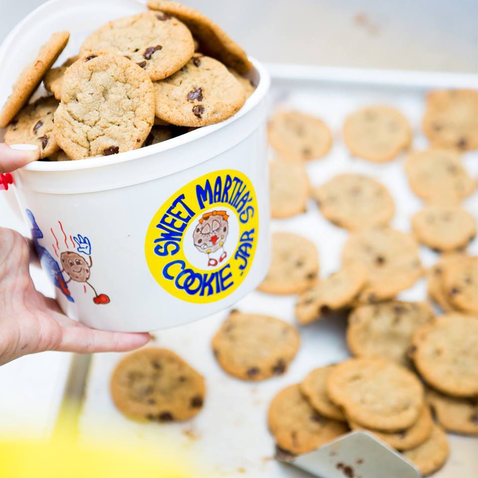 A sheet pan full of chocolate chip cookies with a plastic bucket stacked with cookies in the foreground, held by a hand with red fingernail polish