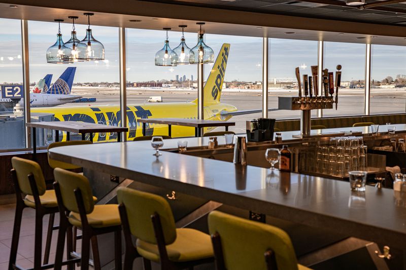 A photo of the bar at People’s Organic. The corner of a bar is set with yellow bar stools; through large windows a large yellow Spirit plane and the tarmac are visible. 