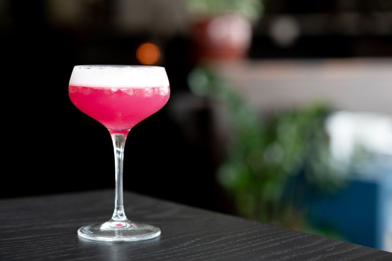 In a coup glass a bright, deep pink cocktail with a white frothy head sits on a black table with a black background. It seems as though there is a spotlight on the drink