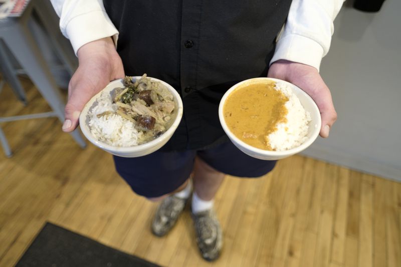 Two hands of a person wearing a black shirt and shorts hold two bowls of food: one is a chicken curry dish with rice, and the other is an orange curry dish with rice. 
