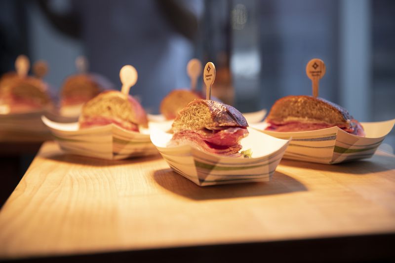 Three half-sandwiches filled with Italian meats in paper boats sit on a wood board underneath a warming light. 