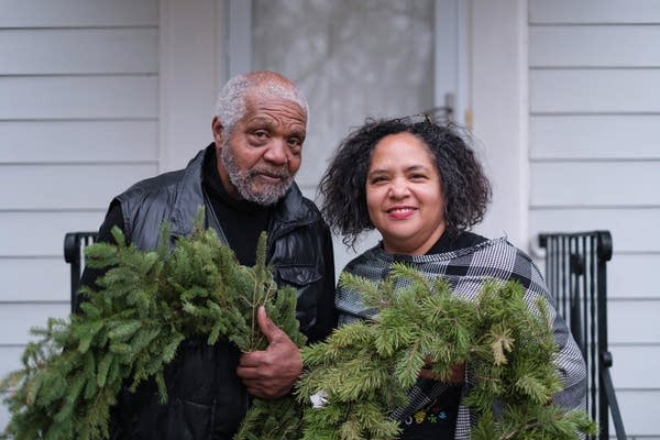 Two people stand in front of a house holding wreaths