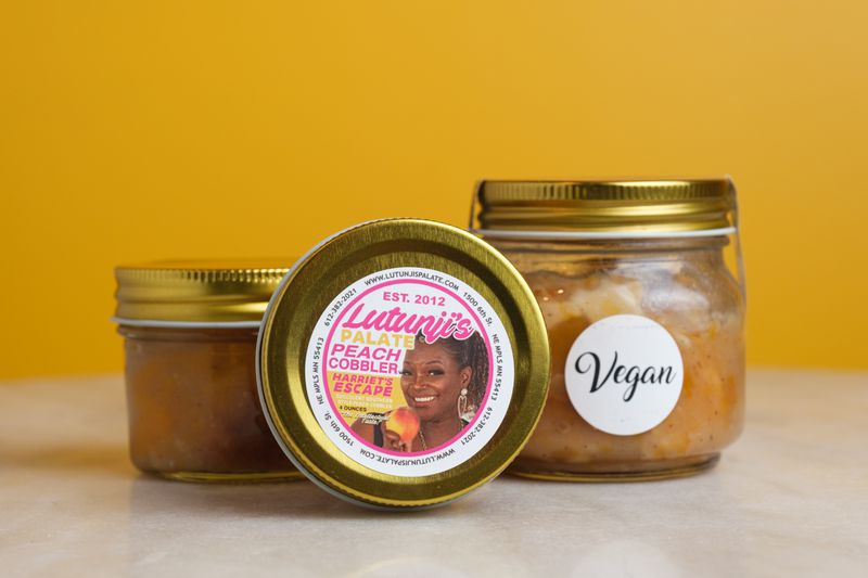 Three jars of peach cobbler sit on a marble table. Two are standing upright, one of them marked with a sticker reading “vegan.” One is balanced on its side with the label facing out. The label shows Lutunji’s face and features pink and orange writing. 