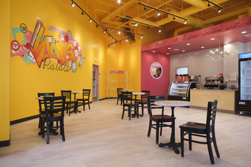 The bakery space has bright yellow and pink walls, and hand-painted mural reading “Lutunji’s Palate.” There are five small round tables with black chairs in the cafe space; in the background, a counter and refrigerators are visible. 