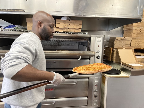 Pulling a pizza out of the over at Rock City Pizza, owner Joseph Charles says one way to offset rising costs of food and labor is to put in more hours himself.