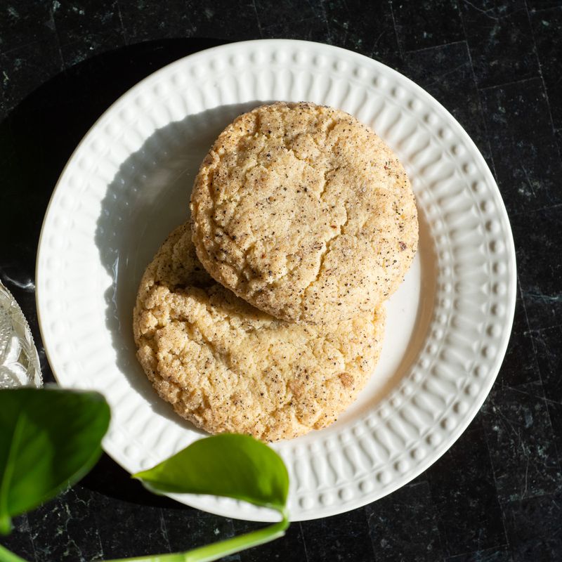 Two Chai snickerdoodles, beige-colored cookies flecked with brown, sit on a white plate on a black background. A green plant is visible in the foreground. 