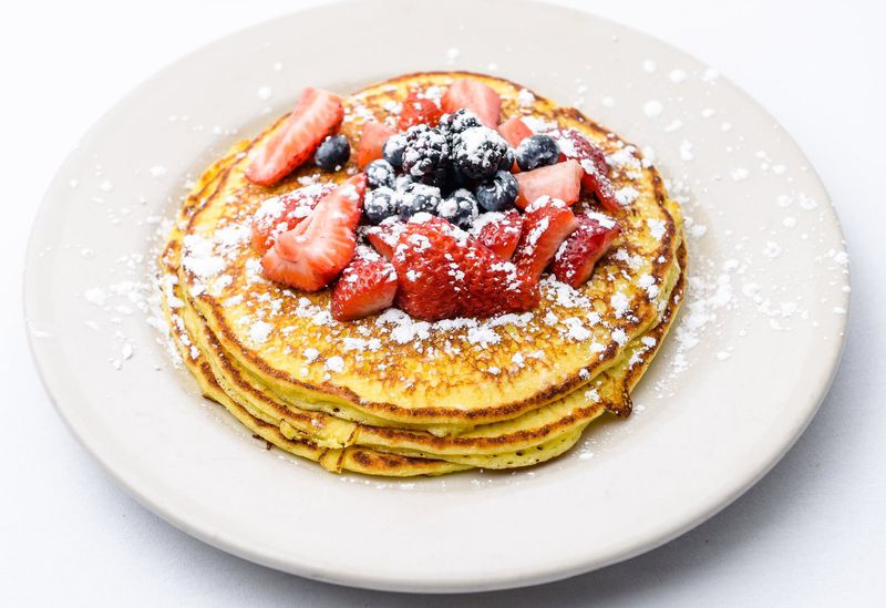 A white plate with a stack of three lemon ricotta pancakes dusted with powdered sugar and topped with strawberries and blueberries on a white background. 