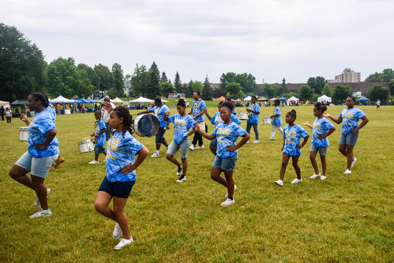 A group of Black girls wearing blue tie-dyed T-shirts march across the grass in the park; in the background are people playing drums and an array of tents. 