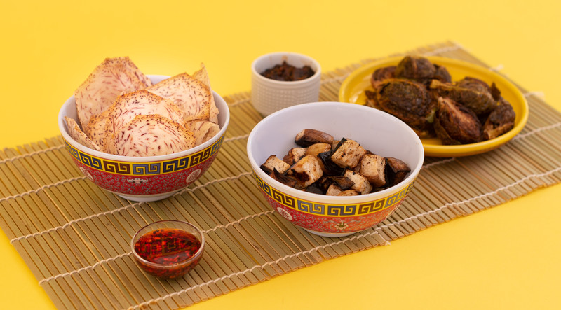 A bowl of tofu, a bowl of taro chips, a plate of barbecue, and a small bowl of chili paste are positioned on a reed mat on a bright yellow background. 