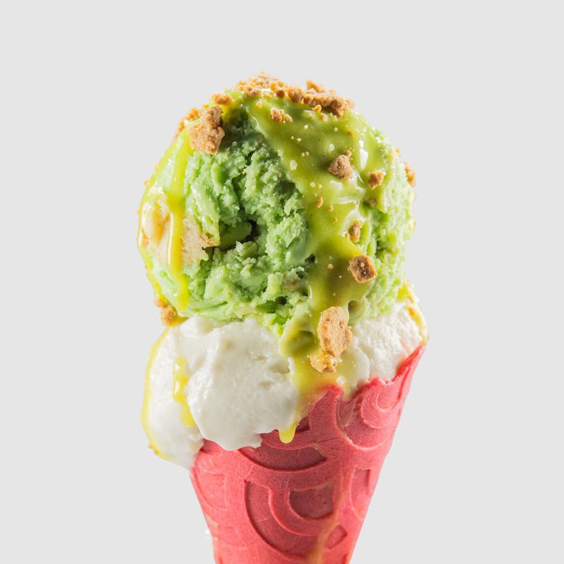 A double scoop ice cream cone with a green ooze dripping over a vanilla scoop to a red waffle cone, with crumbles on top.