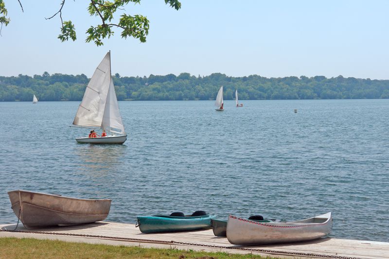 In the foreground, a small sailboat with white sailed helmed by three people in red life jackets floats on a calm lake. In the background, there’s a shoreline of green trees, and three other similar sailboats in the distance. 