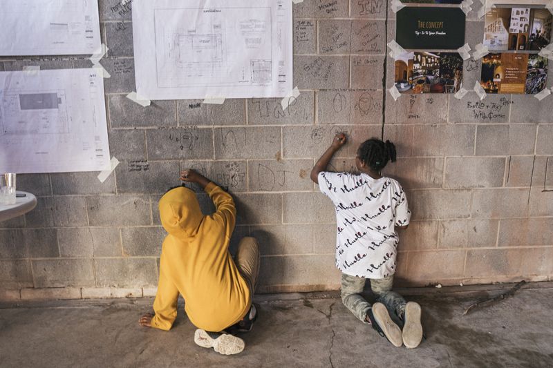Two children, one wearing a yellow sweatshirt and another wearing a white T-shirt with black lettering, kneel on the ground. They are drawing on a gray brick wall that has posters taped to it. 
