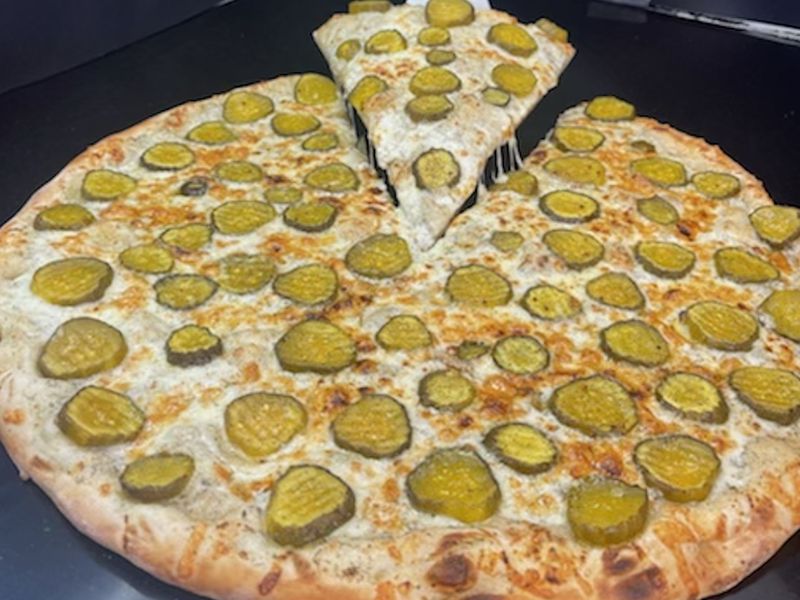 A large round cheese pizza with slices of dill pickle on it; one slice is being lifted out. 