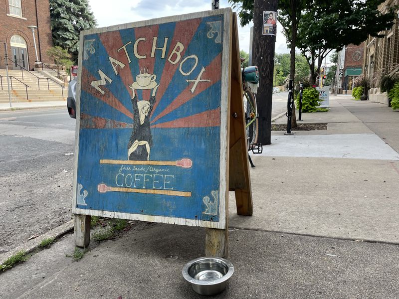 A blue, red, and white wooden sign reading “Matchbox Coffee” with an illustration of a person holding a mug of coffee over their head, on a city sidewalk. 