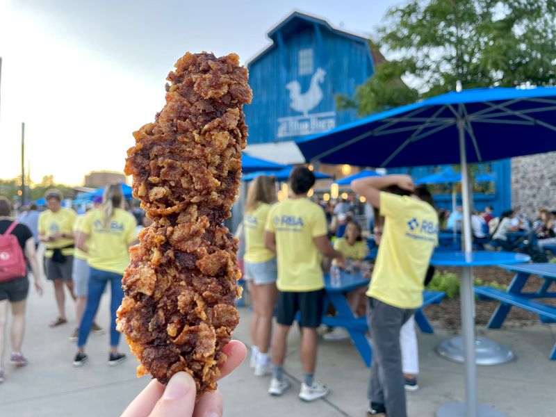 A hand holding a crusted hot chicken tender with a blue barn and people visible in the background. 