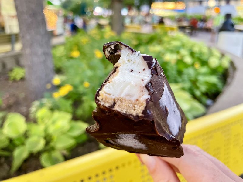 A hand holds a piece of key lime pie dipped in dark chocolate.  A yellow bench and plants are visible in the background. 
