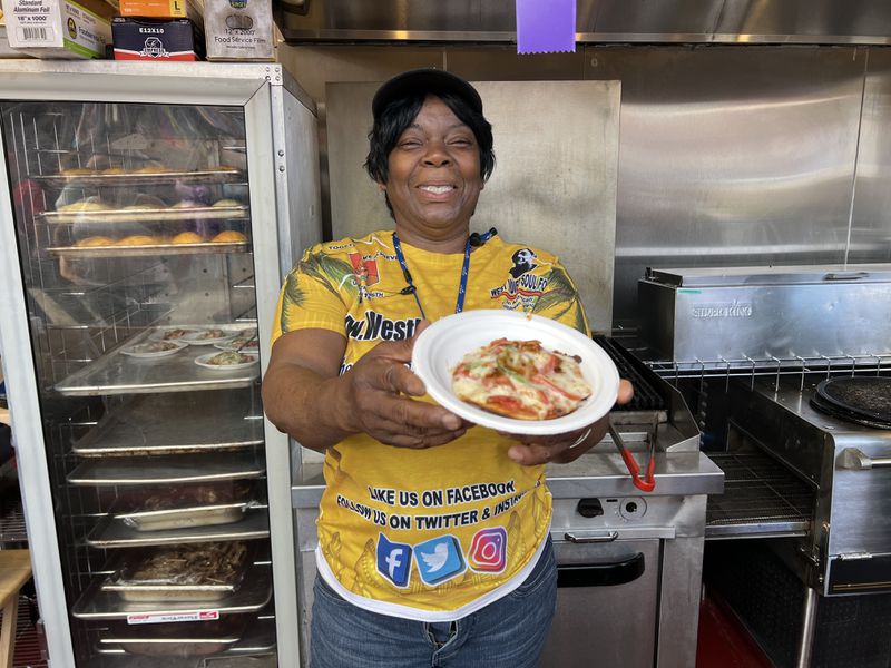 A woman wearing a yellow shirt smiles and holds out a mini pizza on a white plate. 