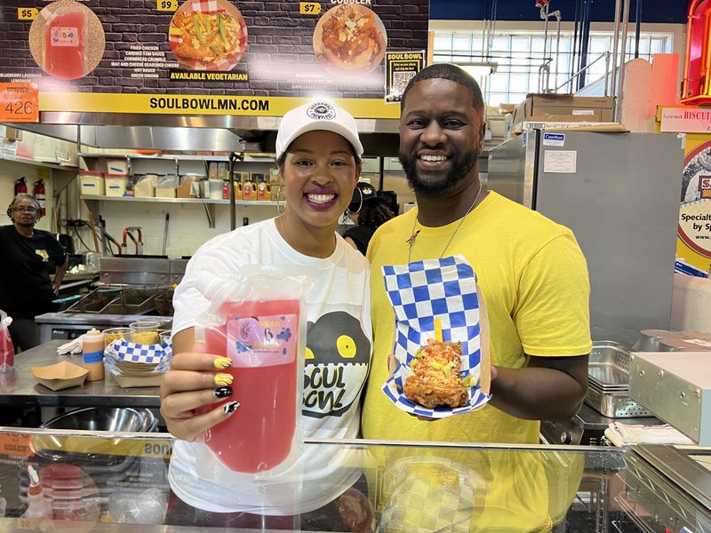 Two people holding fried chicken in a basket and a pouch of pink lemonade smile and pose in a commercial food stand. 