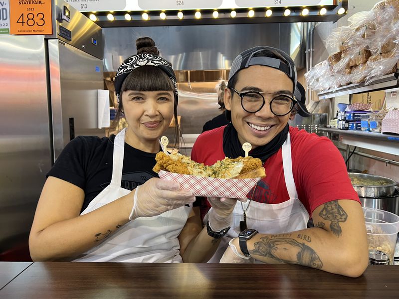 Two people wearing white aprons and standing at a food counter smile for the camera and hold a dish with fried vegan chicken. 