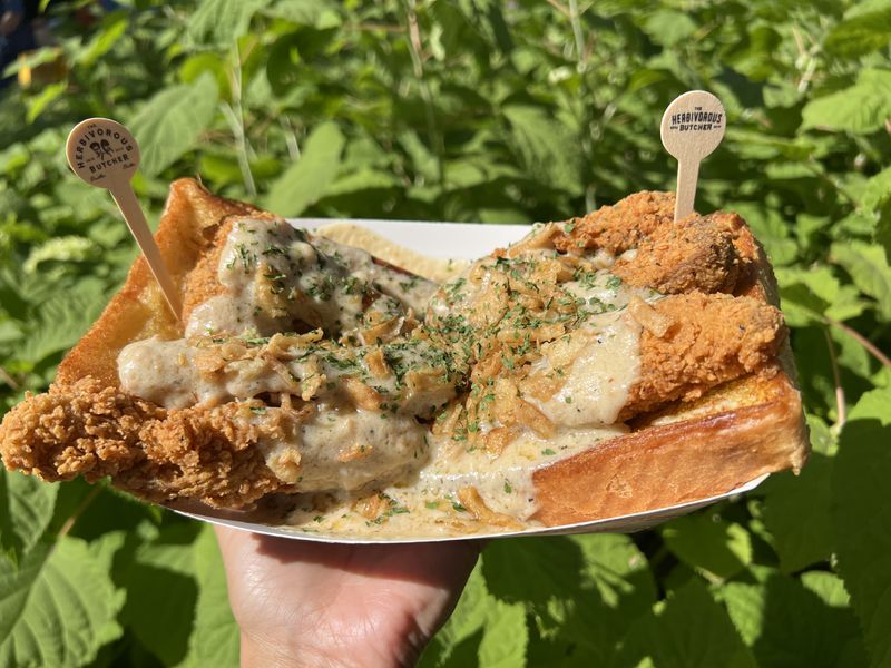 A hand holds a dish of vegan fried chicken on Texas toast in front of a green bush. 